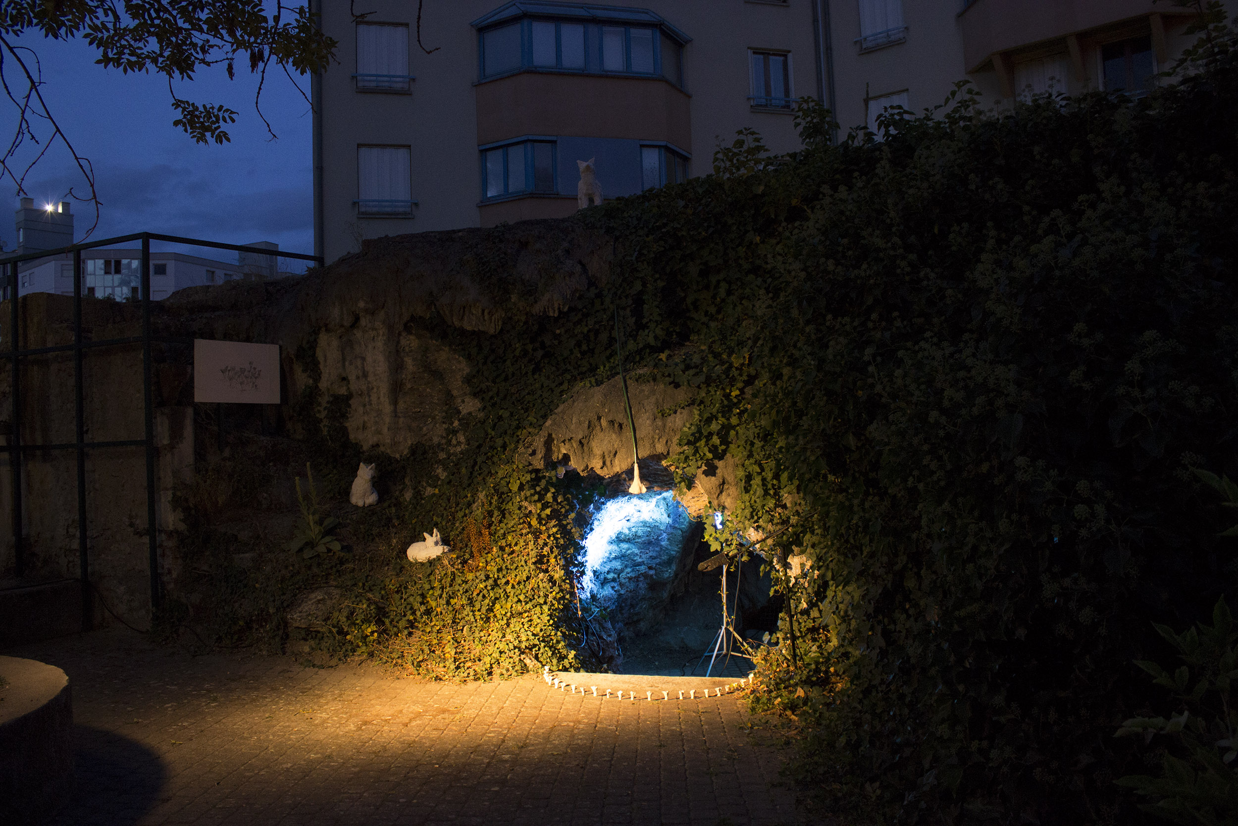 La photo est prise de nuit, on voit l'entrée de la grotte qui est éclérée en bleu depuis l'intérieur. Un spot jaune délimite l'entrée de la grotte. On voit le clercle de chanterelles, 2 renardeaux et le dessin de chanterelles.